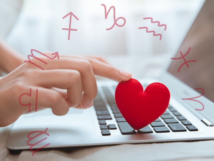 Eine Hand tippt auf eine Computertastatur, während sie ein kleines, rotes Stoffherz berührt. Um die Tastatur und die Hand schweben Symbole der Tierkreiszeichen. Das Bild symbolisiert digitale Kommunikation oder Online-Dating im Zusammenhang mit Astrologie und Liebe. Der Hintergrund ist weich und verschwommen, was eine intime und moderne Atmosphäre vermittelt.