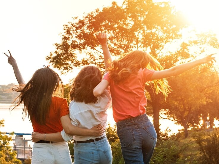 Drei junge Frauen stehen Arm in Arm mit dem Rücken zur Kamera, während sie die Arme freudig in die Luft werfen. Sie stehen in einer sonnigen Landschaft, umgeben von Bäumen, mit Blick auf einen Fluss oder See im Hintergrund. Die Sonne scheint warm und strahlend, was eine lebendige und fröhliche Atmosphäre schafft.