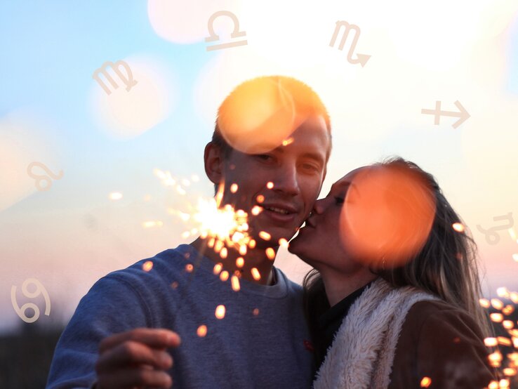 Das Bild zeigt ein Pärchen in einer romantischen Stimmung bei Sonnenuntergang. Die Frau gibt dem Mann einen Kuss auf die Wange, während er eine brennende Wunderkerze in der Hand hält. Im Hintergrund leuchten helle Lichtreflexe, die eine warme, festliche Atmosphäre erzeugen. Um die beiden herum sind Symbole der Sternzeichen wie Waage, Löwe und Skorpion zu sehen, was auf ein astrologisches Thema hinweist. Die Szene wirkt intim und fröhlich und vermittelt eine stimmungsvolle, romantische Stimmung.