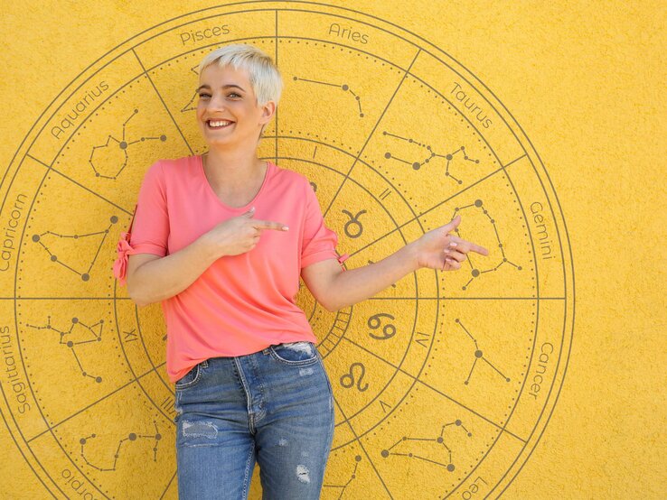 Eine lächelnde Frau mit kurzem, blondem Haar steht vor einer gelben Wand, auf der ein astrologisches Rad mit den Tierkreiszeichen abgebildet ist. Sie trägt ein pinkfarbenes T-Shirt und blaue Jeans und zeigt mit beiden Händen auf den Bereich des Horoskops. Die Szene wirkt freundlich und einladend, mit einem sonnigen Hintergrund, der die fröhliche Stimmung unterstreicht.