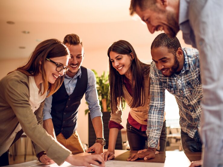 Eine Gruppe von fünf jungen Erwachsenen arbeitet gemeinsam an einem Tisch in einem modernen Büro. Sie sind in ein kreatives Projekt vertieft, lachen und zeigen Begeisterung. Eine Frau mit Brille und einem beigen Blazer zeigt auf ein Dokument oder eine Skizze, während die anderen aufmerksam zusehen. Die Gruppe ist vielfältig, mit verschiedenen Stilen und Hintergründen. Das warme Licht im Raum verleiht der Szene eine freundliche und motivierende Atmosphäre, die Zusammenarbeit und Teamgeist symbolisiert.