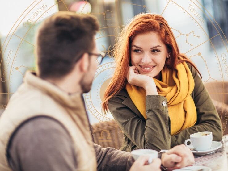 Ein junges Paar sitzt in einem Café und unterhält sich bei einer Tasse Kaffee. Die Frau mit roten Haaren und gelbem Schal lächelt ihren Gesprächspartner an, während sie ihren Kopf auf eine Hand stützt. Im Hintergrund ist ein astrologisches Rad mit den zwölf Tierkreiszeichen und Sternbildern eingeblendet, was eine Verbindung zwischen dem Gespräch und astrologischen Einflüssen andeutet. Die Szene wirkt warm und harmonisch.