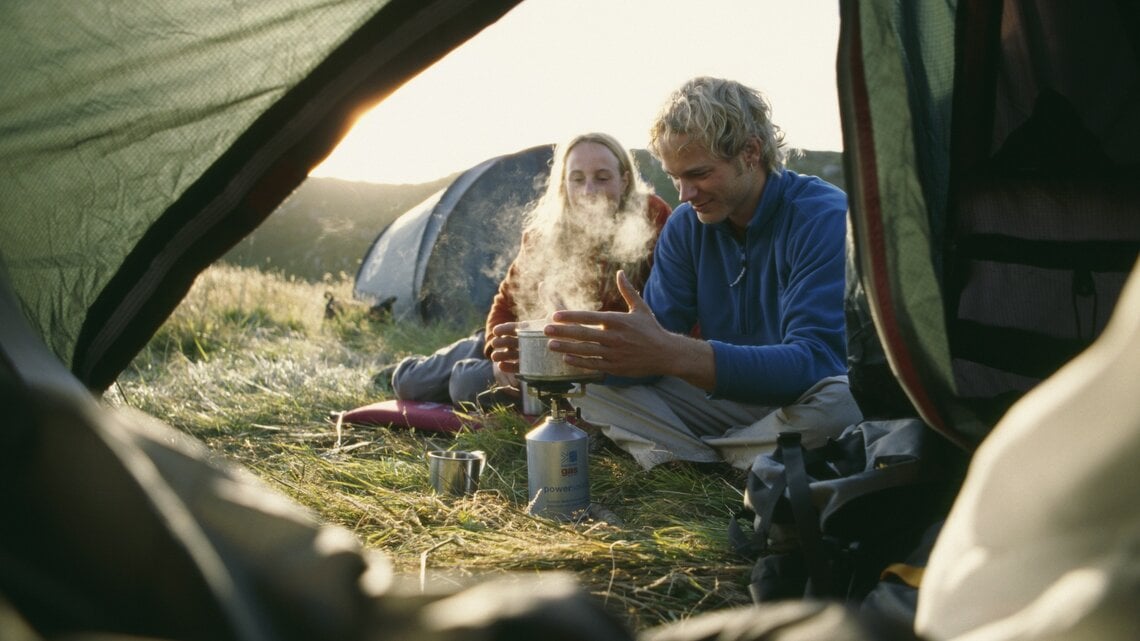 Ein Paar beim Zelten sitzt vor einem Gaskocher, auf dem etwas heiß aufkocht. Im Hintergrund geht die Sonne auf. 