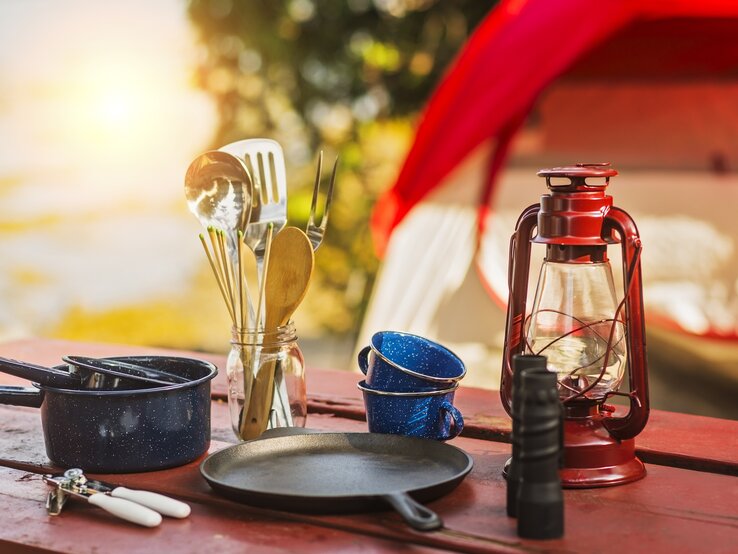 Campinggeschirr und Kochutensilien auf einem roten Tisch, daneben eine rote Laterne und ein blaues Emaille-Set im Sonnenuntergang. | © Getty Images/Daniel Grill/Tetra Images