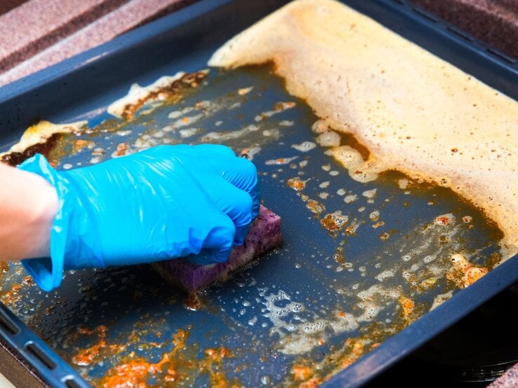 Eine Hand mit blauen Gummihandschuhen schrubbt mit einem Schwamm ein schmutziges Backblech mit Seifenlauge.