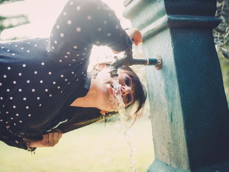 Nahaufnahme einer Frau in gepunkteter Bluse, die sich über einen Wasserhahn im Park beugt und trinkt.