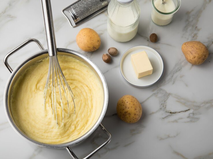 Kartoffelpüree in einem Topf mit Schneebesen, daneben Kartoffeln, Butter, Milch und Muskatnüsse auf hellem Marmortisch.