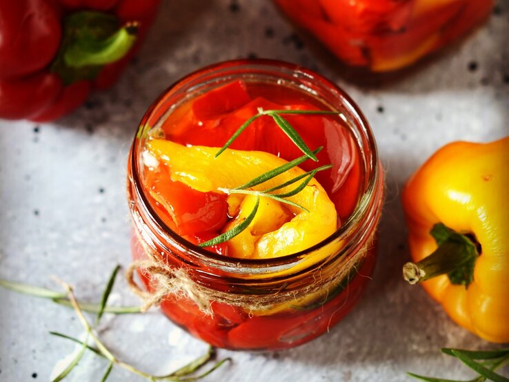 Eingelegte Paprika in einem Glas auf einem grauen Steintisch mit Kräutern und Olivenöl. Alles von schräg oben fotografiert.