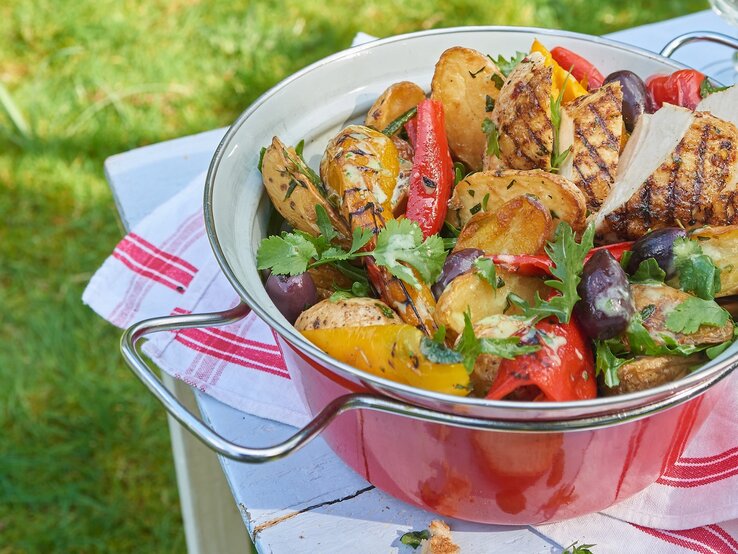 Eine rote Schüssel mit dem Grillkartoffelsalat mit Hähnchen. Alles auf einem Tisch mit weißer Tischdecke und rötlicher Deko