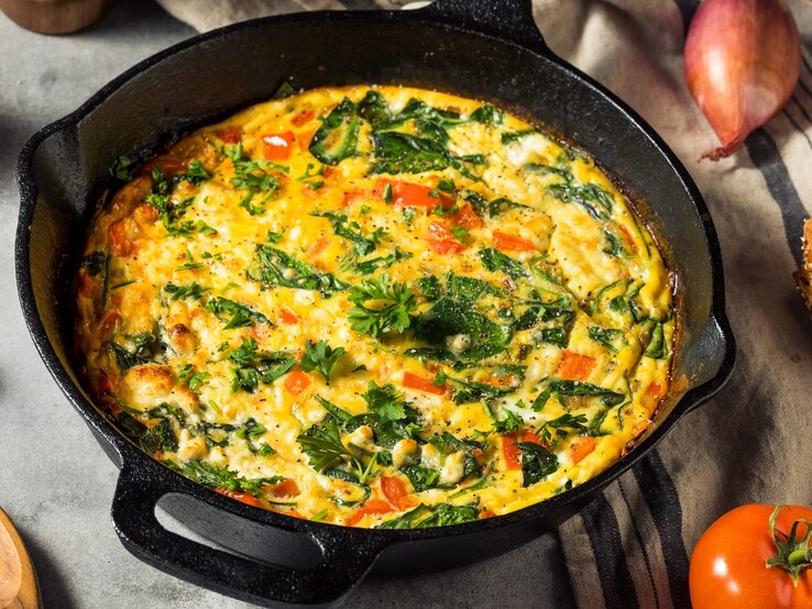 Eine gusseiserne Pfanne mit dem Omelette mit Feta und Tomaten auf einem grauen Steintisch. Alles von schräg oben fotografiert.