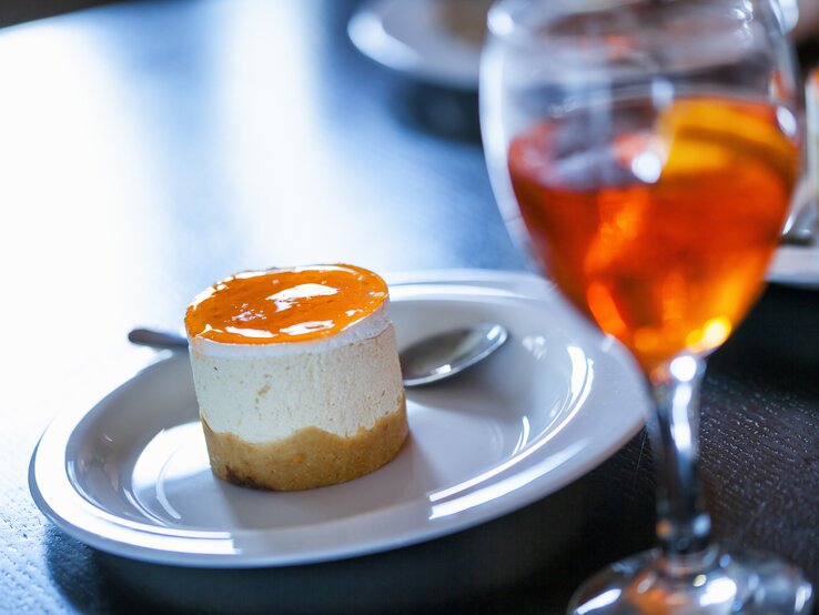 Ein kleiner Aperol-Käsekuchen mit einem orangenen Guss aus Orangensaft, Prosecco und Aperol neben einem Glas mit Aperol Spitz