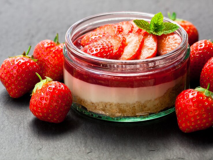 Frisch zubereitetes Erdbeerdessert im Glas mit Keksboden, Sahneschicht und Erdbeerscheiben, umgeben von frischen Erdbeeren auf dunklem Hintergrund.