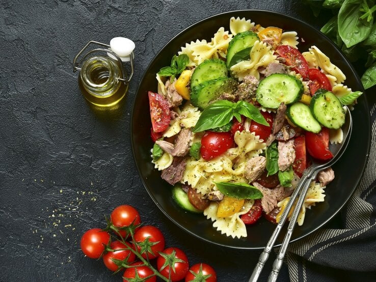 Ein Teller mit Nudelsalat aus Farfalle, Tomaten, Gurken und Thunfisch auf dunklem Untergrund, daneben eine Flasche Olivenöl und Kirschtomaten.