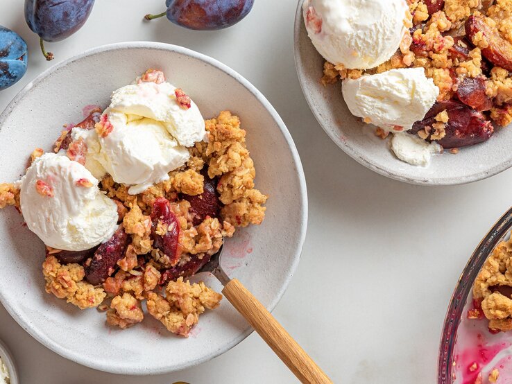 Zwei Teller mit dem Pflaumen-Crumble und Vanilleeis auf einem hellen Tisch mit weiteren frischen Pflaumen drum herum, direkt von oben fotografiert.