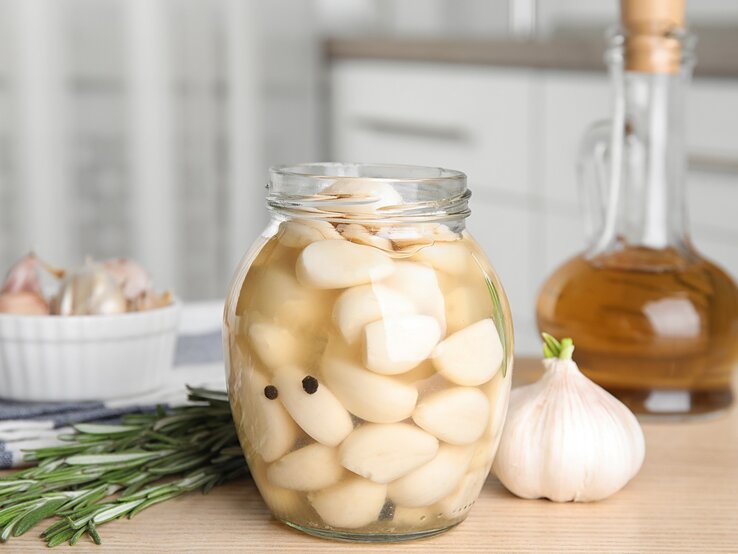 In einem hellen Küchenumfeld stehen ein Glas mit eingelegten Knoblauchzehen und eine Knoblauchknolle neben Rosmarin und Ölflasche.