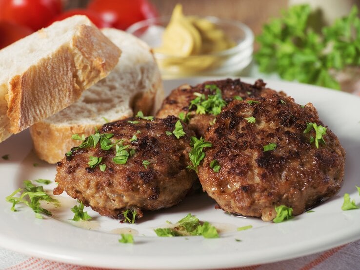 Mehrere Frikadellen bestreut mit Kräutern auf weißem Teller neben Brotscheiben. 