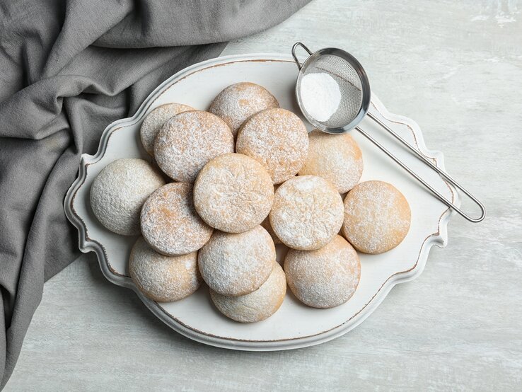 Mehrere mit Puderzucker bestäubte Butterwölkchen-Plätzchen auf weißem Servierteller.