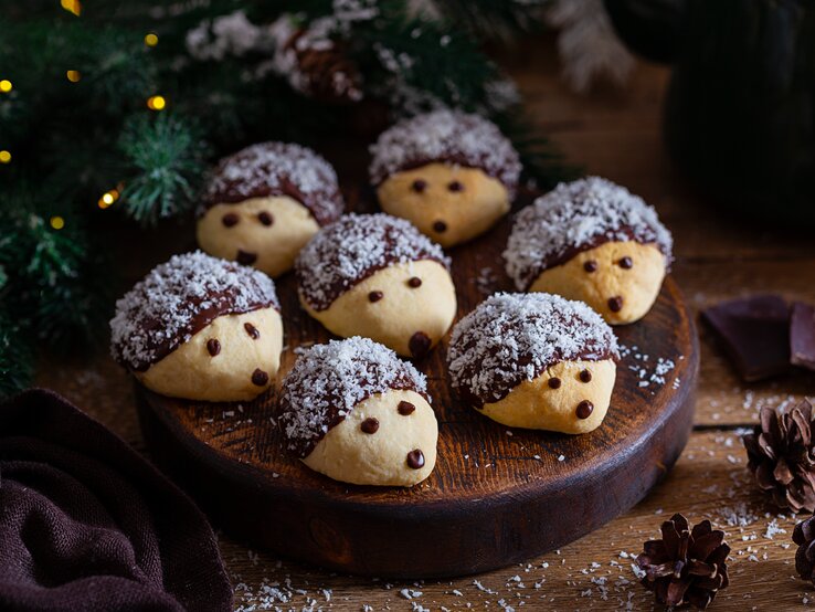 Mehrere mit Kokosraspel dekorierte Igelkekse auf einem Holzbrett in weihnachtlichem Ambiente. 