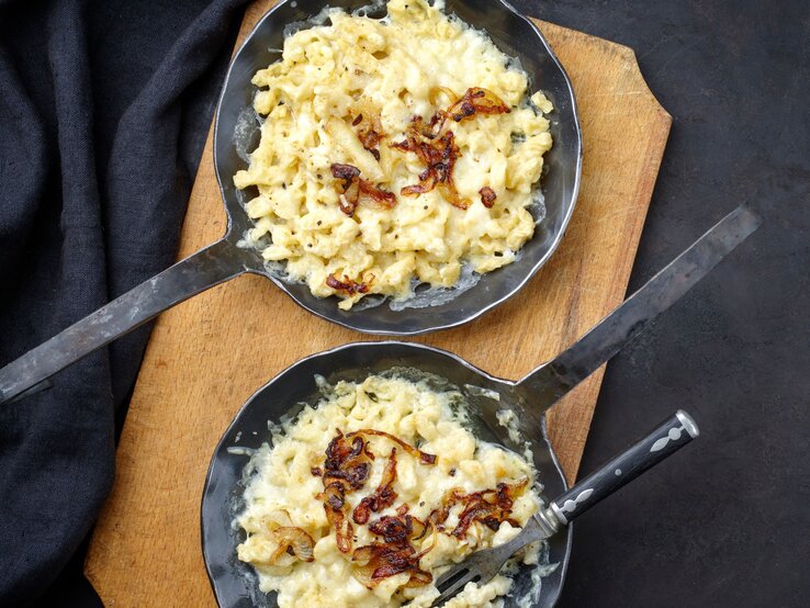 zwei gusseiserne Pfannen gefüllt mit Käsesätzle, garniert mit gebratenen Zwiebeln auf einer dunklen Tischdecke. 