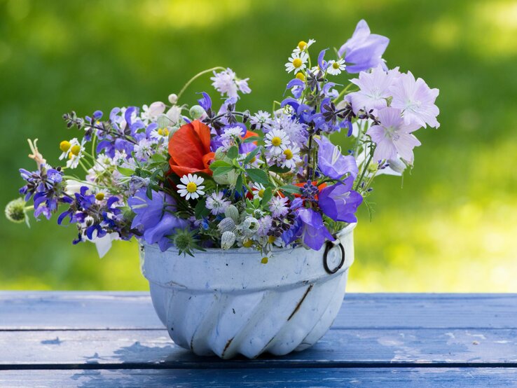 Farbenfrohe Wildblumen in einer alten, weißen Backform auf einem blauen Holztisch vor grünem Hintergrund.