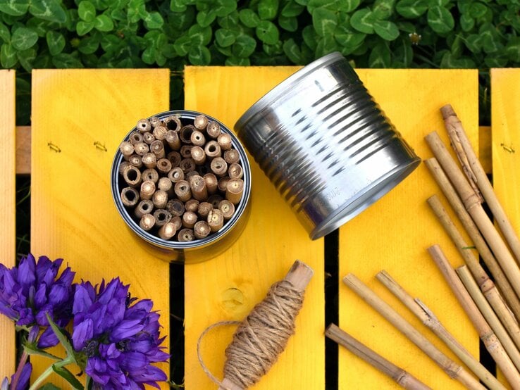 Bunte Insektenhotels in einer Blechdose, daneben eine umgestürzte Dose und Holzstäbe auf gelbem Holztisch mit lila Blumen.