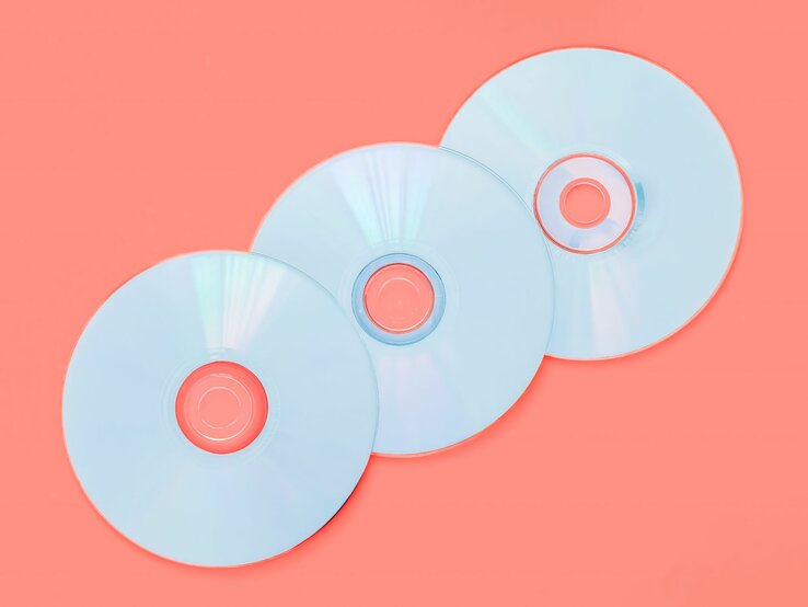Drei silberne CDs liegen auf einem korallenroten Hintergrund, mit leichtem Schimmer von Lichtreflexen.