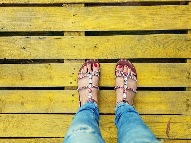 Eine Person mit roten Fußnägeln trägt funkelnde Sandalen und steht auf gelben Holzplanken.