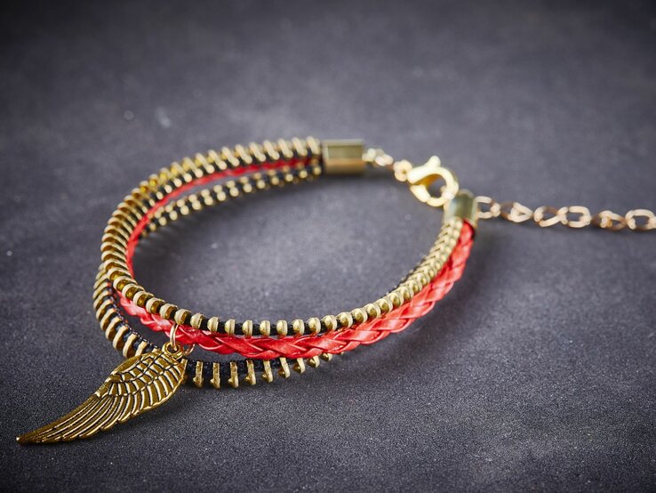 Rotes geflochtenes Lederarmband mit goldener Engelsflügel-Anhänger und Reißverschluss auf schwarzem Hintergrund.