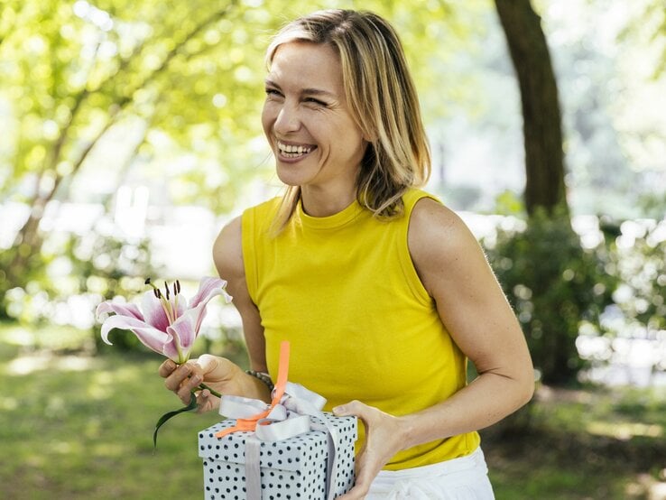 Eine blonde Frau in einem gelben Top hält lächelnd eine rosa Lilie und eine Explosionsbox im sonnigen Park.