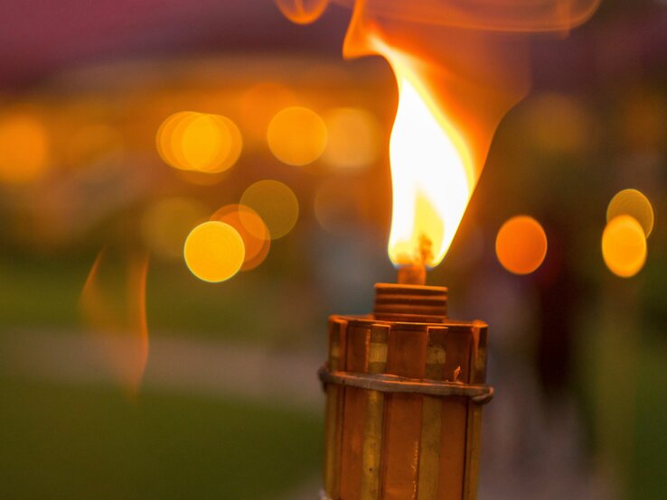 Eine brennende Fackel im Vordergrund mit unscharfen, warmen Lichtern im Hintergrund in der Abenddämmerung.