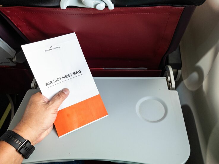 Eine Hand hält einen Spuckbeutel über einem heruntergeklappten Tablett in einem Flugzeug mit dunkelrotem Rücksitz.