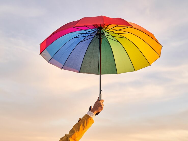 Hand in gelber Jacke hält einen bunten Regenschirm vor einem pastellfarbenen Himmel am Abend.