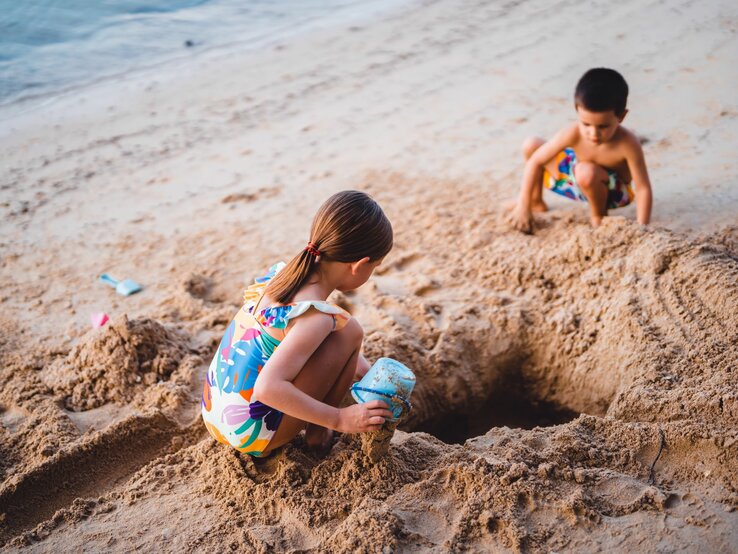 Zwei junge Kinder in farbenfroher Strandkleidung buddeln im Sand an einem leeren Strand ein Loch.