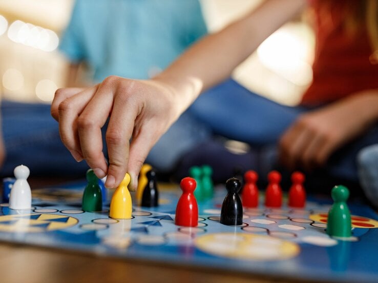 Nahaufnahme einer Hand, die eine gelbe Spielfigur auf einem Brettspiel zieht, unscharfer Hintergrund mit spielenden Kindern.