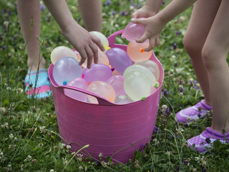 Zwei Kinder beugen sich über einen rosa Eimer voller bunter Wasserbomben auf einer grünen Wiese.