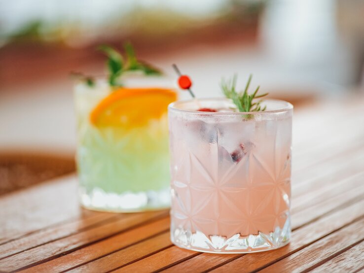 Zwei erfrischende Cocktails in Kristallgläsern auf einem Holztisch, der vordere rosa mit Rosmarin und Beeren, der hintere grün mit Orangenscheibe und Minze.