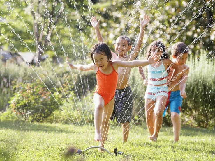 Vier Kinder in bunter Badebekleidung rennen lachend durch Wasserstrahlen eines Rasensprengers auf einer grünen Wiese an einem sonnigen Tag.