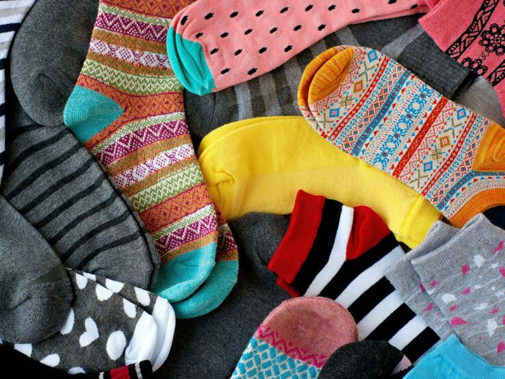 Bunte Socken mit verschiedenen Mustern und Farben liegen chaotisch auf einem grauen Untergrund.