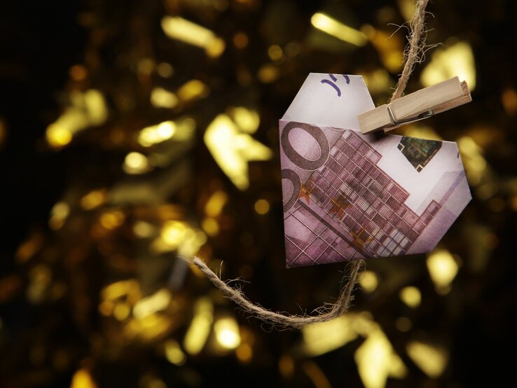 Ein Herz aus einem 200-Euro-Schein hängt an einer Wäscheklammer vor einem goldenen, unscharfen Hintergrund.