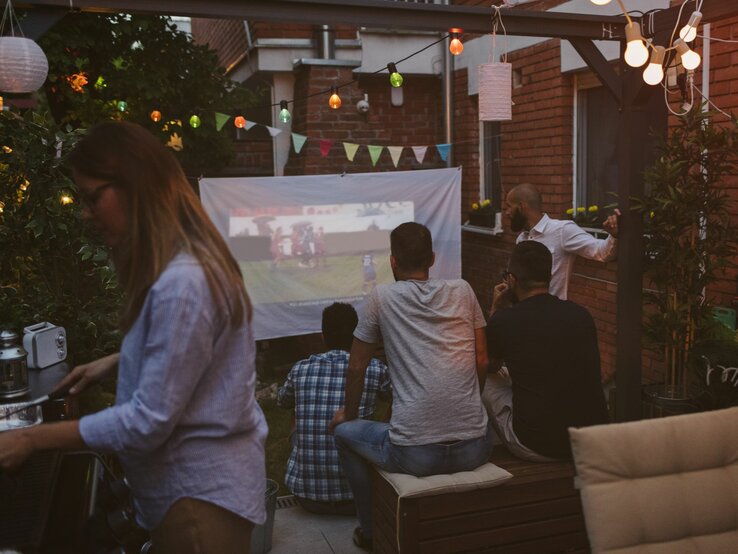 Eine Gruppe von Männern schaut am Abend im Garten ein Fußballspiel auf einer Leinwand, während eine Frau am Grill steht.