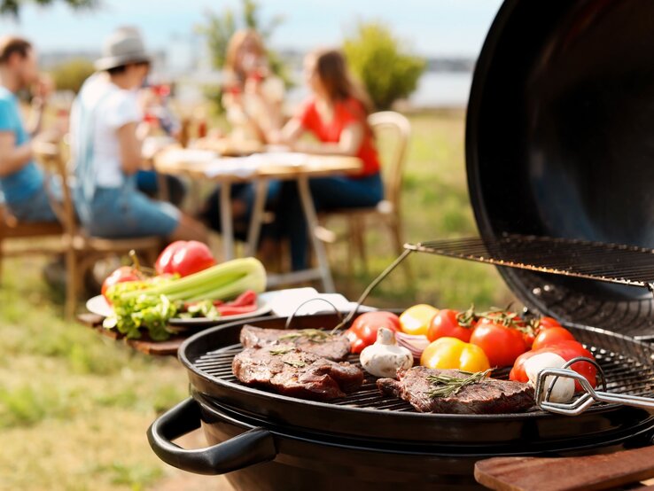 Bunte Gemüse und Steaks werden auf einem Grill zubereitet, während eine Gruppe von Freunden im Hintergrund lacht und isst.