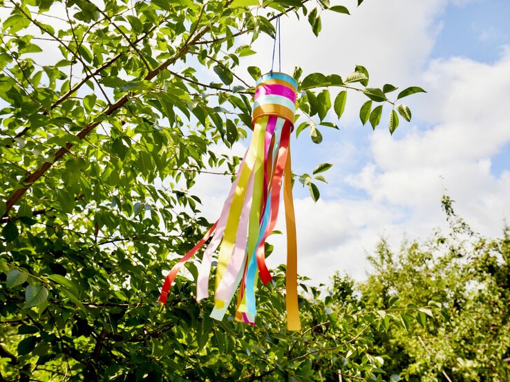 Ein Windspiel aus bunten Bändern weht im Sommerwind an einem grünen Ast unter einem klaren, bewölkten Himmel.