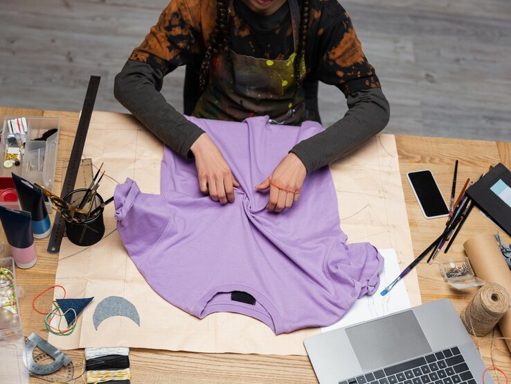 Kreative Arbeit an einem lila T-Shirt, umgeben von Pinsel, Lineal, Laptop und Bastelmaterialien auf einem Holztisch.