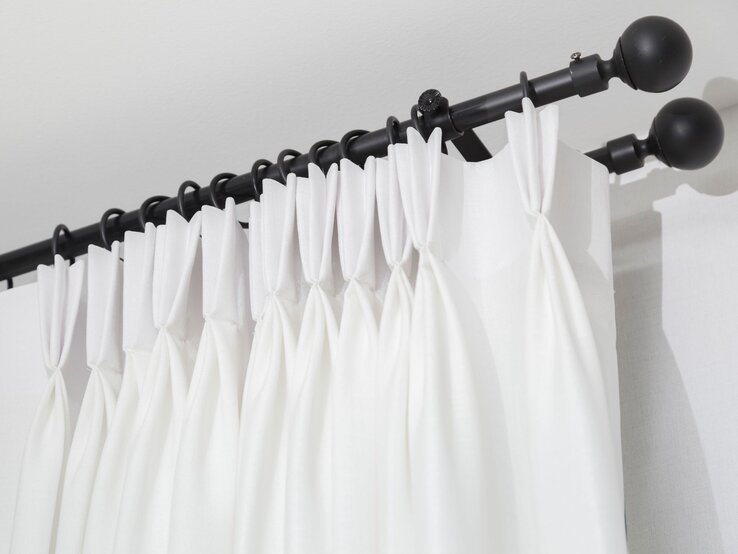 Ein weißer Vorhang mit Falten hängt mithilfe von Gardinenringen an einer schwarzen Gardinenstange mit Kugelenden vor einer weißen Wand.