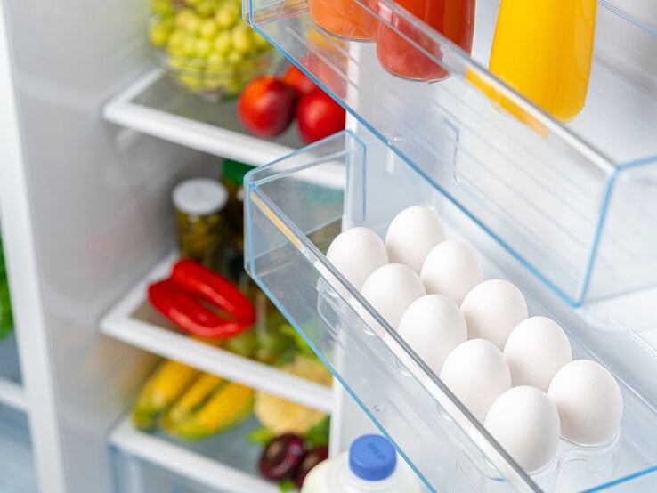 Ein geöffneter Kühlschrank zeigt Eier, Säfte und frisches Gemüse wie Paprika und Mais in den Regalen.
