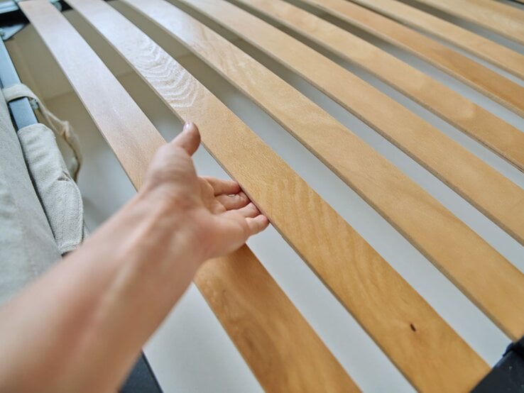 Eine Hand greift nach einer Holzlatte in einem Lattenrost, seitlich ein graues Textilband.