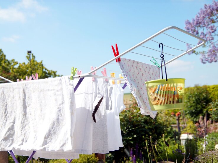 Weiße Wäsche und ein gemustertes Tuch hängen auf einem Wäscheständer im sonnigen Garten mit blühenden Bäumen.