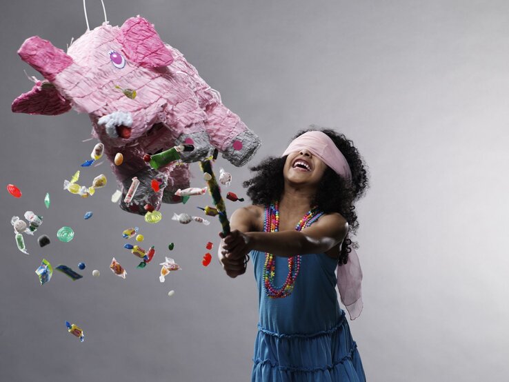 Mädchen mit lockigem Haar und bunter Halskette zerschlägt fröhlich eine rosa Elefanten-Piñata, aus der Bonbons herausfallen.