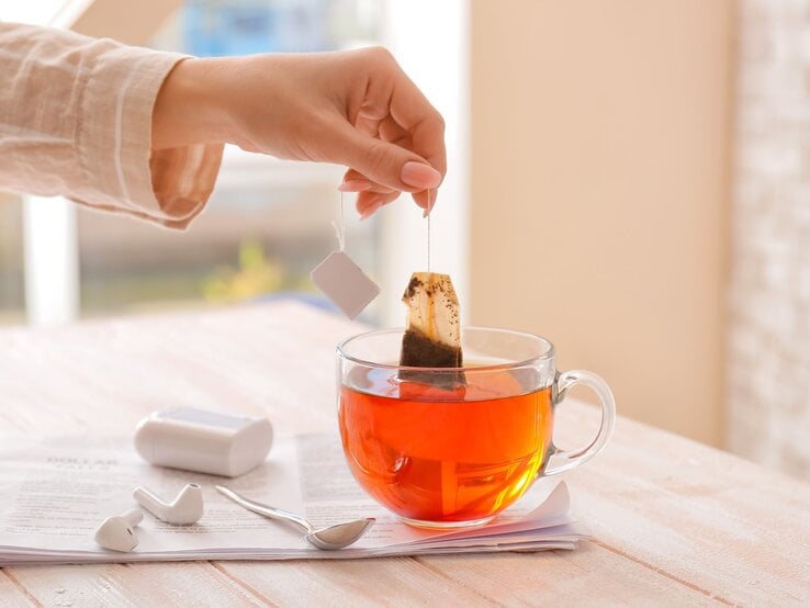 Eine Hand in beiger Bluse taucht einen Teebeutel in eine klare Tasse mit rotem Tee auf einem Holztisch neben weißen Ohrstöpseln und einem Löffel.