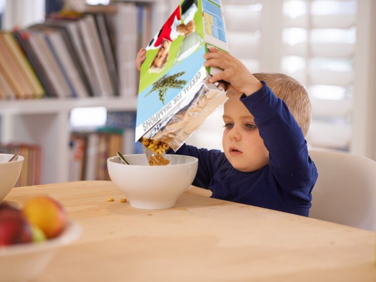 Ein kleiner Junge in blauem Pullover füllt konzentriert Müsli aus einer bunten Packung in eine weiße Schüssel am Holztisch.